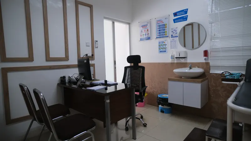 Ruang praktik dokter umum dengan meja kerja dokter, kursi pasien, tempat tidur periksa, dan lemari peralatan medis di area yang sama.
